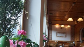 Un restaurant avec des tables et des chaises en bois, une plante en pot aux fleurs roses et un mur de briques orné d’un tableau encadré.
