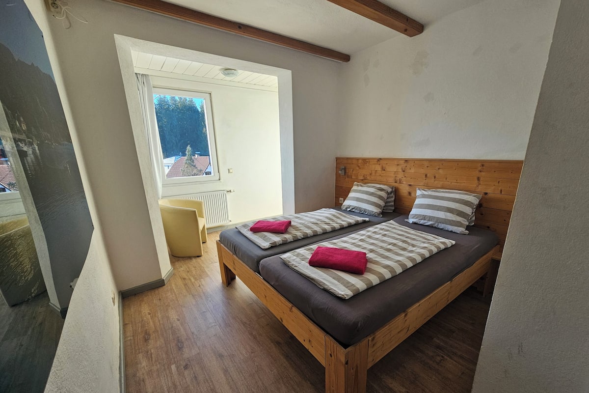 A bedroom with a wooden bed, two pillows, a red towel, and a window with a view of a building.