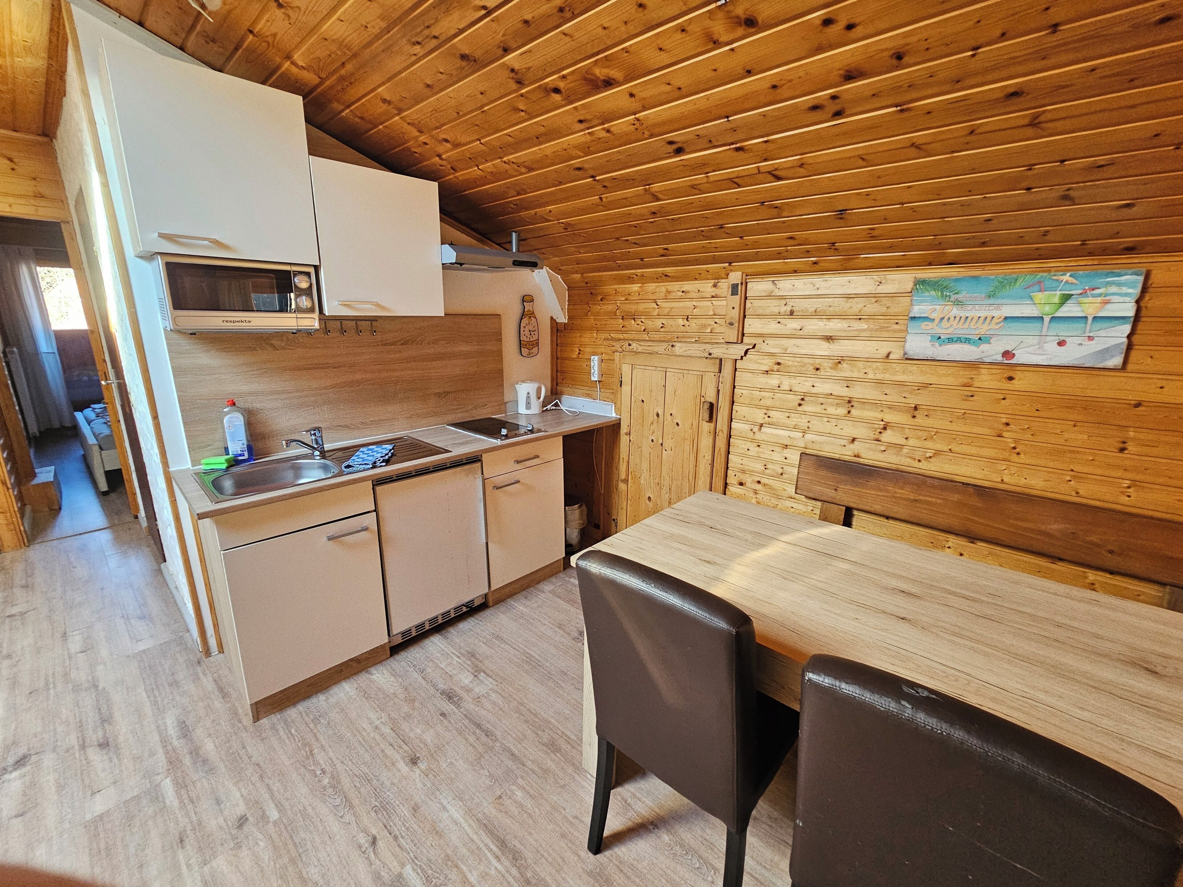 Een kleine keuken met houten kastjes, een spoelbak en een eethoek met een tafel en stoelen.