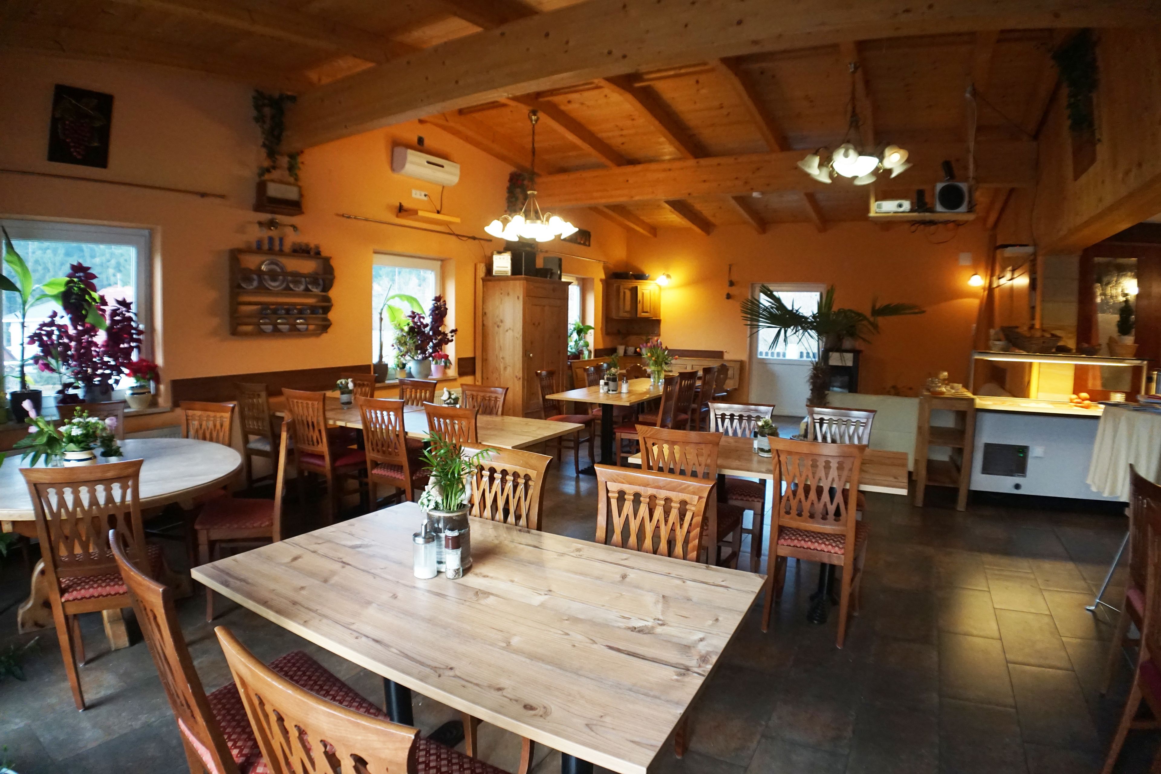 A spacious dining area with wooden tables and chairs, surrounded by potted plants and a high ceiling with exposed beams.