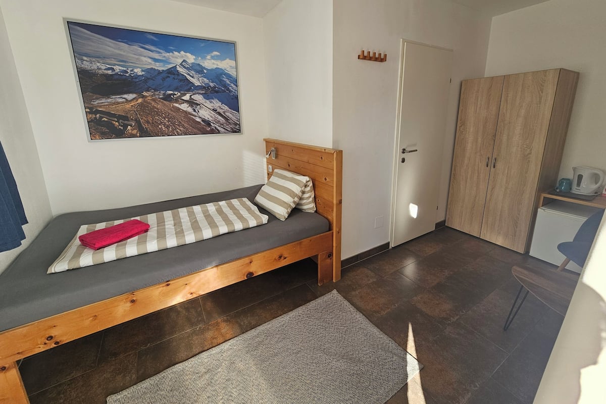 A small, neatly arranged bedroom with a bed, a wooden wardrobe, a door, and a large landscape painting on the wall.