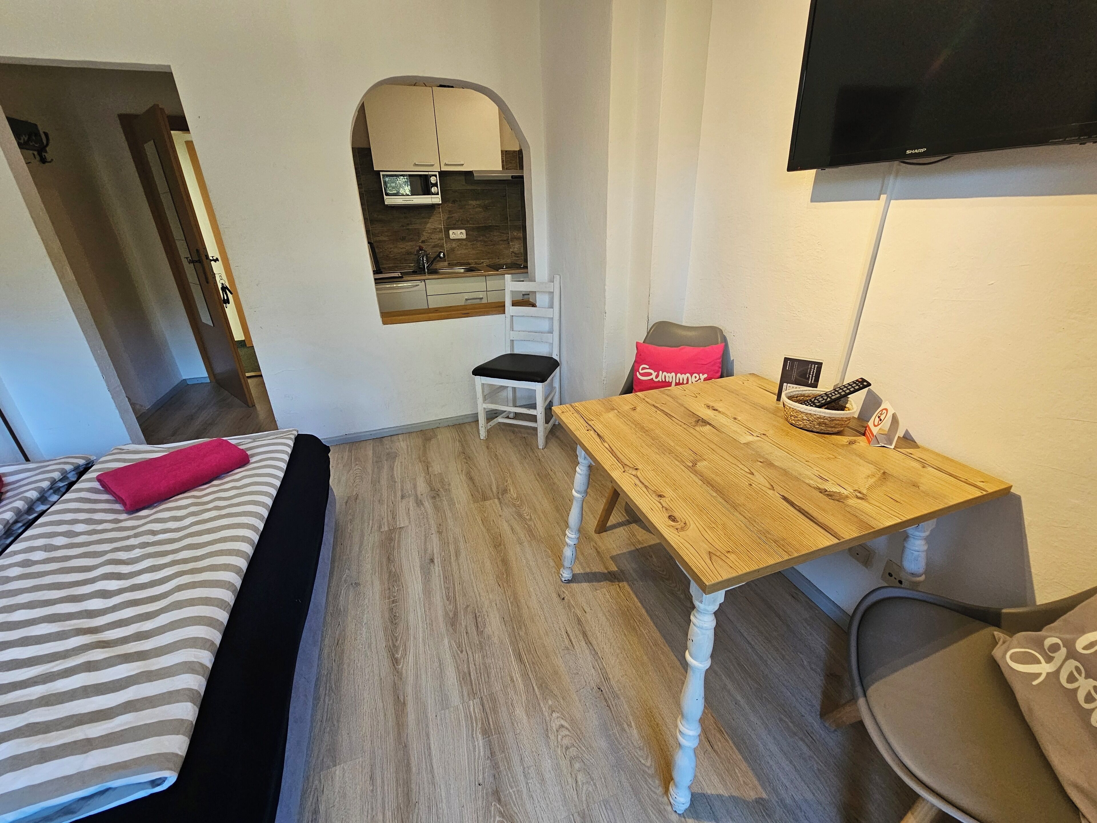A compact room with a bed, a wooden table, a chair, a television, and a kitchenette.