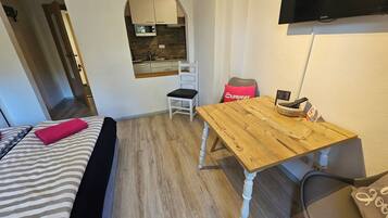 A compact room with a bed, a wooden table, a chair, a television, and a kitchenette.