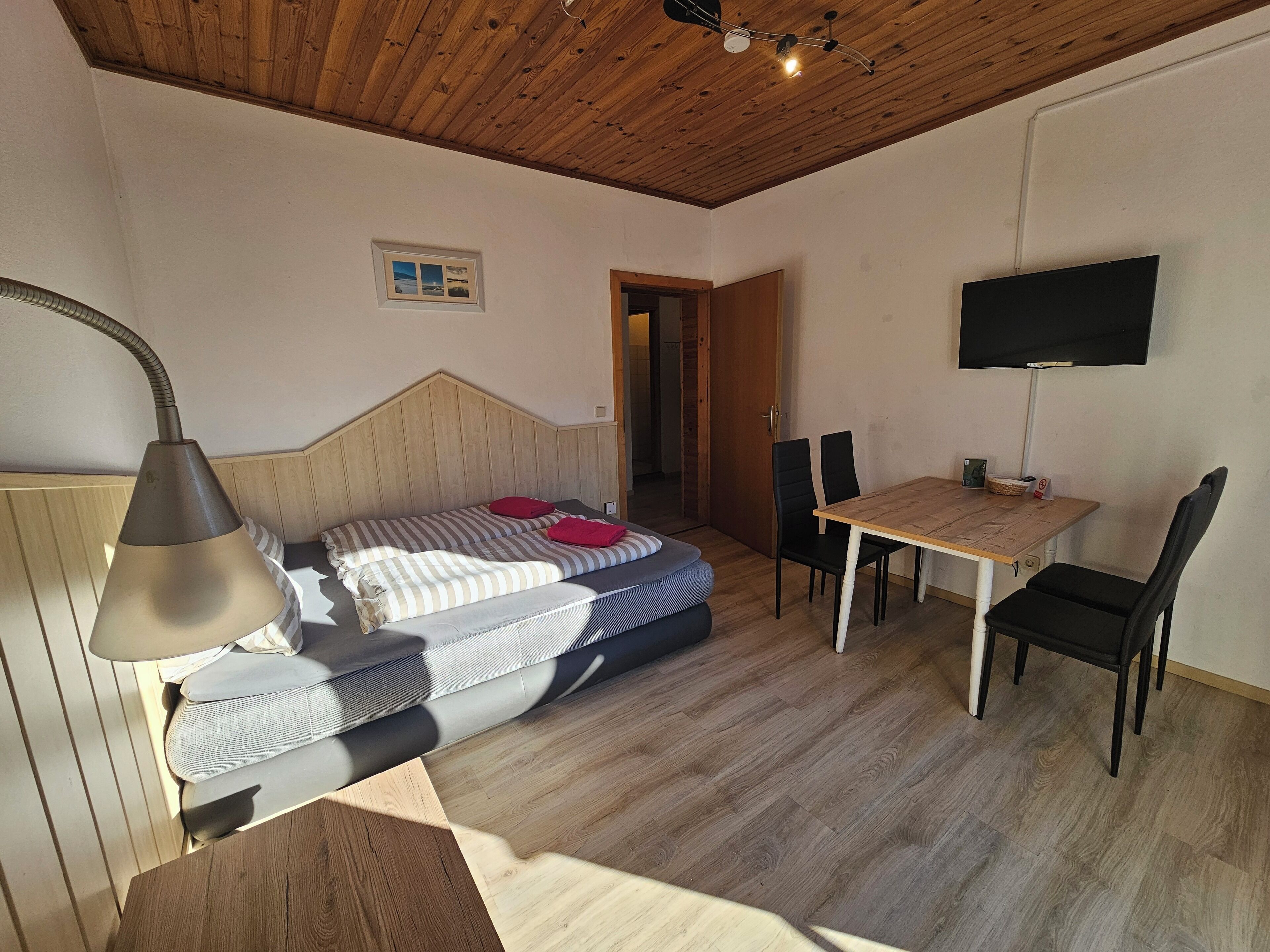 Un dormitorio con techo de madera, una cama con ropa de cama a rayas, una lámpara de noche, una mesita con sillas y un televisor en la pared.