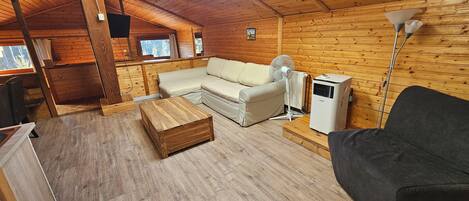 Interior acogedor de una cabaña de madera con sofá, un sillón negro, una mesa de centro de madera, un ventilador de pie y un calentador.