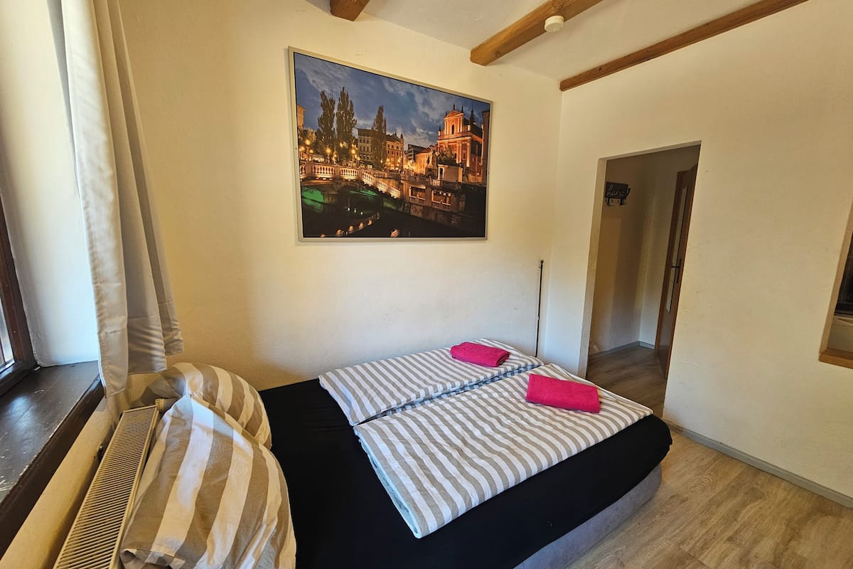 A bedroom with a bed, a radiator, a painting of a cityscape, and a wooden ceiling beam.