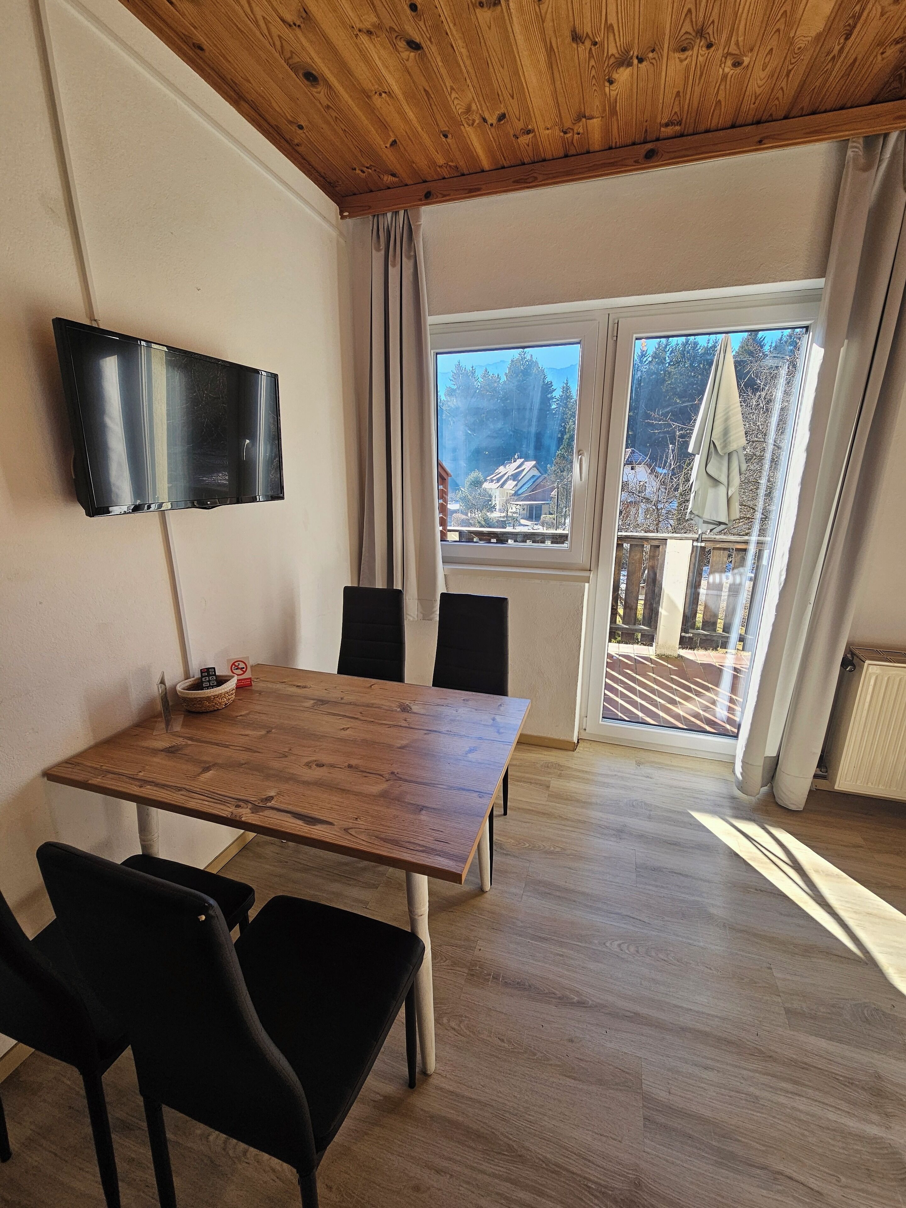 A room with a wooden table, chairs, a flat-screen TV, and a large window with curtains.