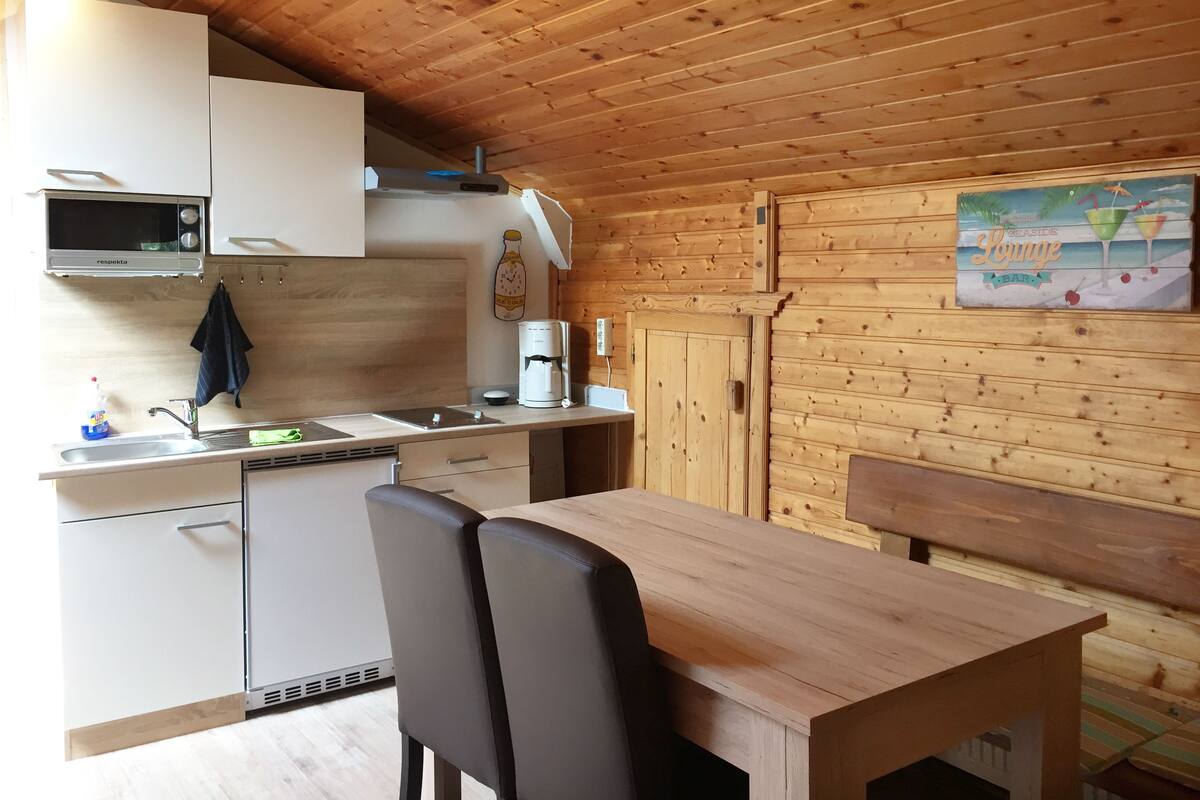 A compact kitchen with a microwave, sink, and coffee maker. There is a wooden table with chairs and a bench. The room has wooden walls and a sloped ceiling.