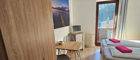 Una stanza con una scrivania in legno, un letto con asciugamani rosa, una TV e un'ampia finestra vista edifici.
