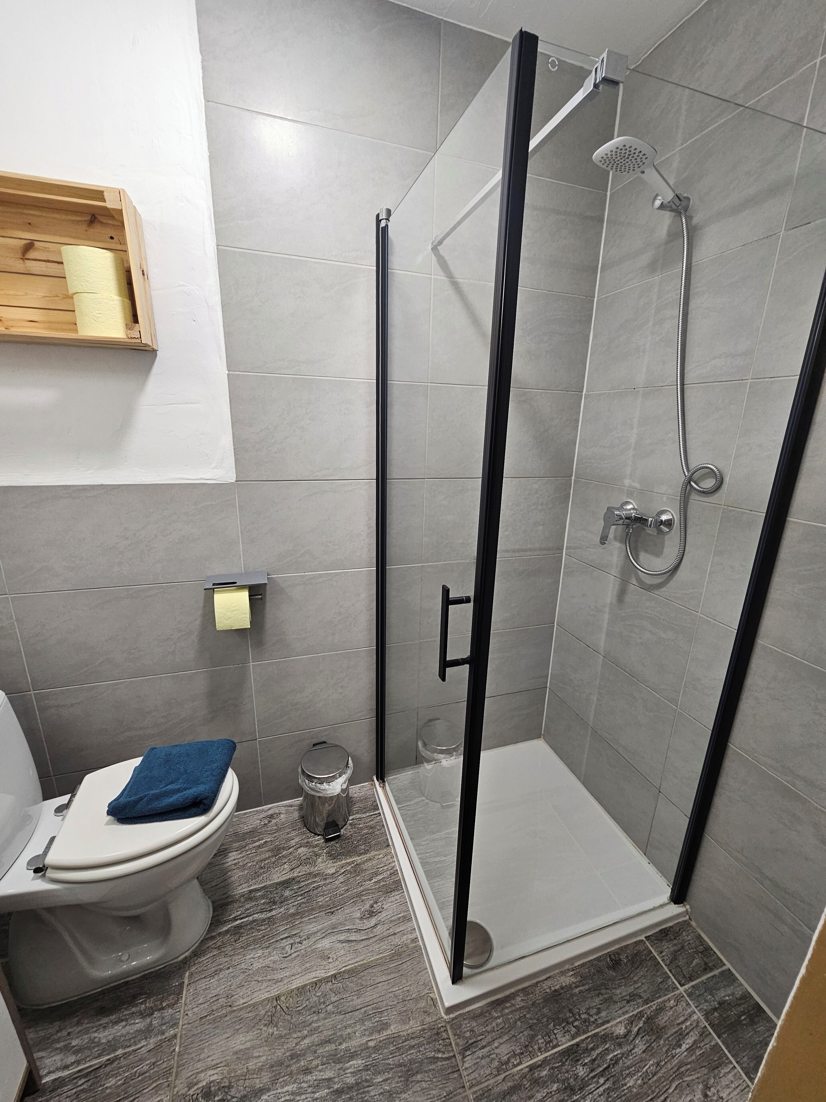 Een moderne badkamer met een douchecabine van glas, een toilet met een blauwe handdoek en een houten plankje met een rol toiletpapier.