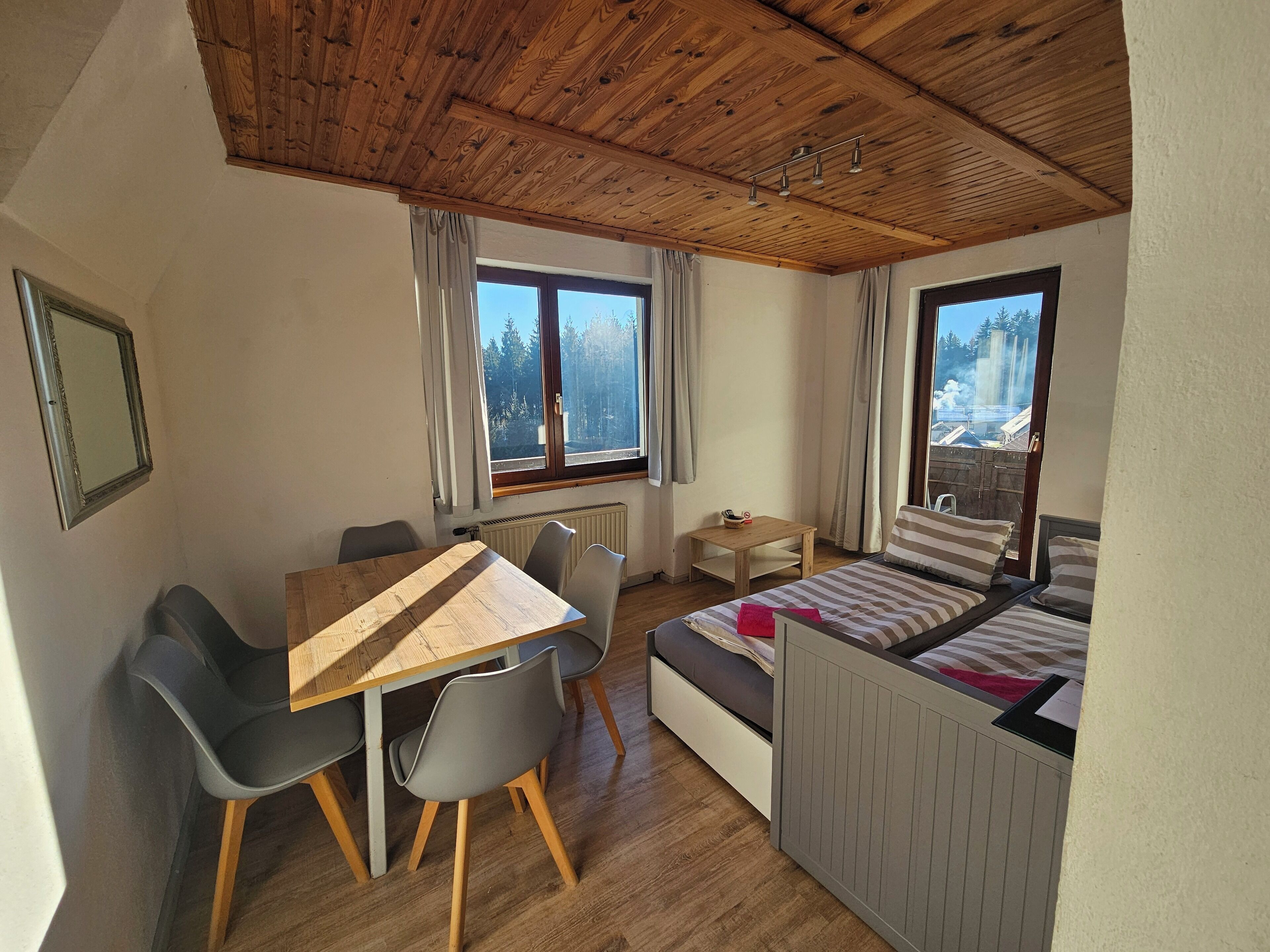 Ein gemütlicher Raum mit Holzbalkendecke, einem Bett mit gestreifter Bettwäsche, einem kleinen Tisch mit Schale, zwei Stühlen und großen Fenstern mit Blick auf Bäume und einen Wasserfall.