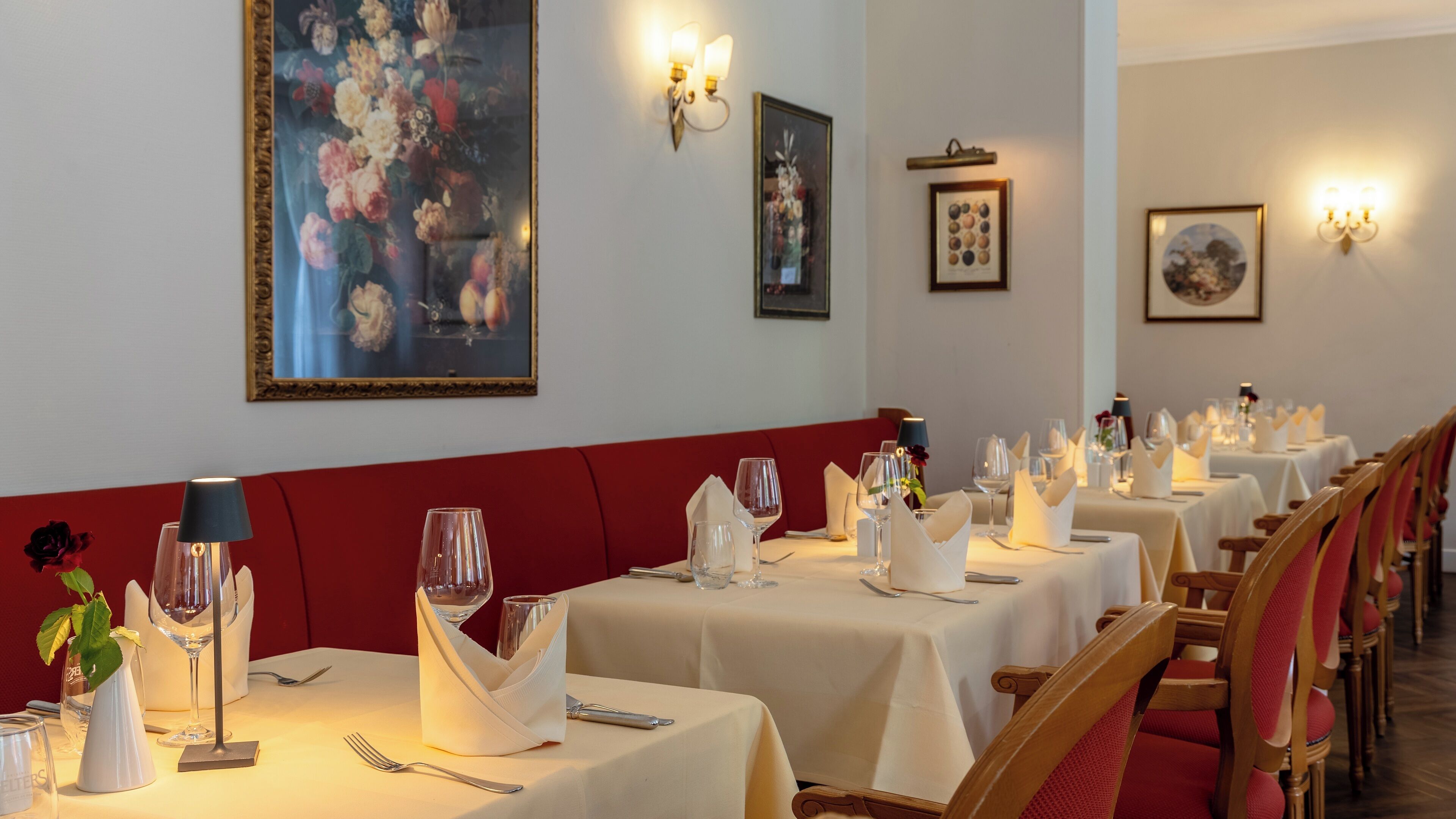 Um restaurante com mesas preparadas para refeições, um grande quadro floral na parede e iluminação embutida.