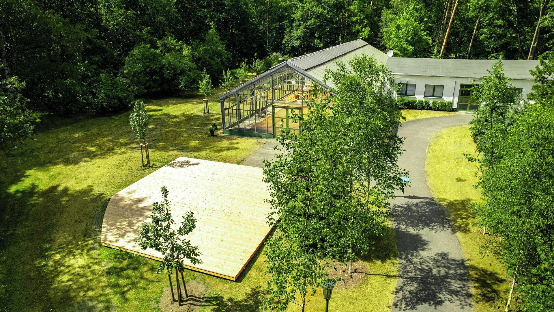 A modern house with a large glass conservatory, surrounded by lush greenery and a paved deck.
