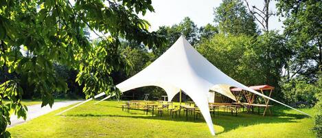 A white tent-like structure with tables and chairs arranged underneath, surrounded by trees and grass.
