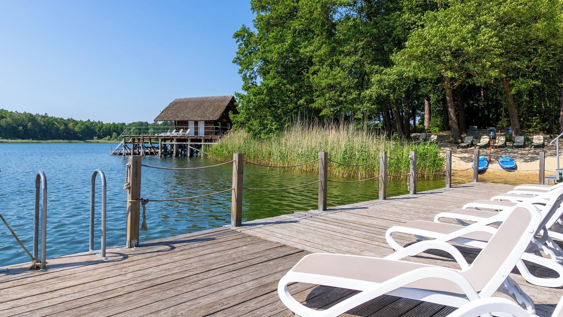A wooden dock with lounge chairs, a thatched-roof building, and a forested shoreline.