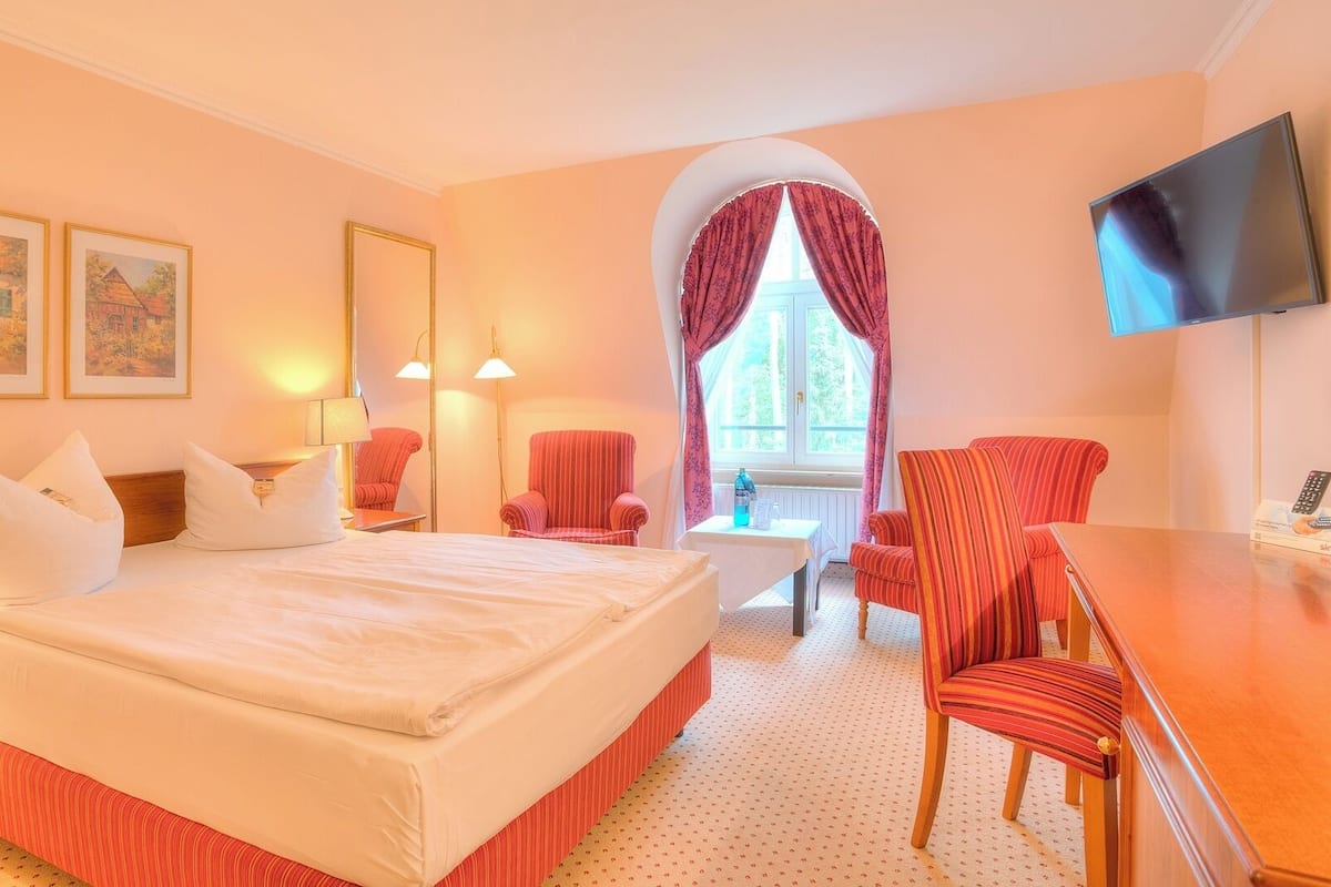 A hotel room with a bed, two chairs, a desk, a TV, and artwork on the walls.