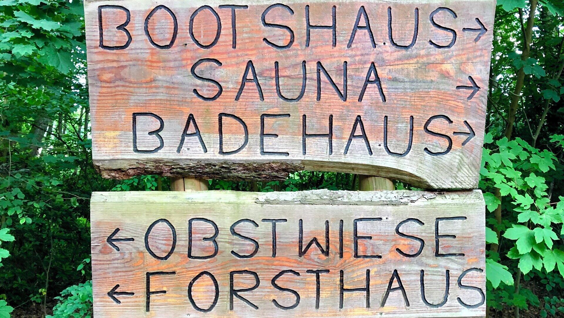 Un letrero de madera con flechas direccionales y texto que indica los nombres de varios establecimientos: BOOTSHAUS, SAUNA, BADEHAUS, OBSTWIESE y FORSTAUS.