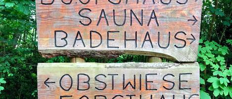 Un letrero de madera con flechas direccionales y texto que indica los nombres de varios establecimientos: BOOTSHAUS, SAUNA, BADEHAUS, OBSTWIESE y FORSTAUS.