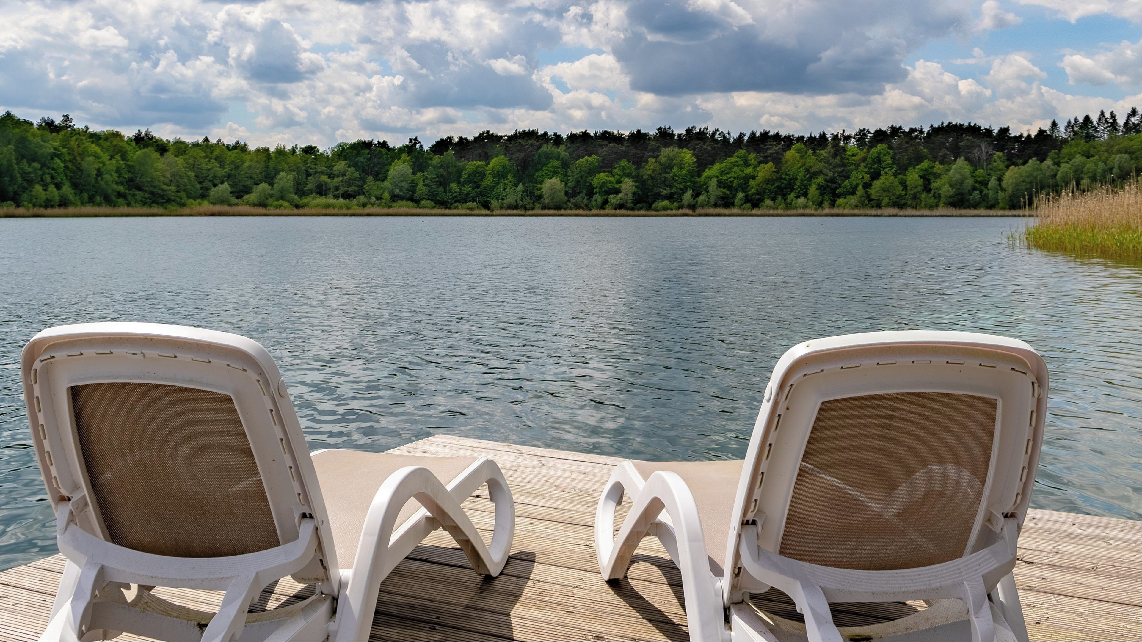 Dos sillas blancas en una terraza de madera con vistas a un lago y una orilla boscosa.