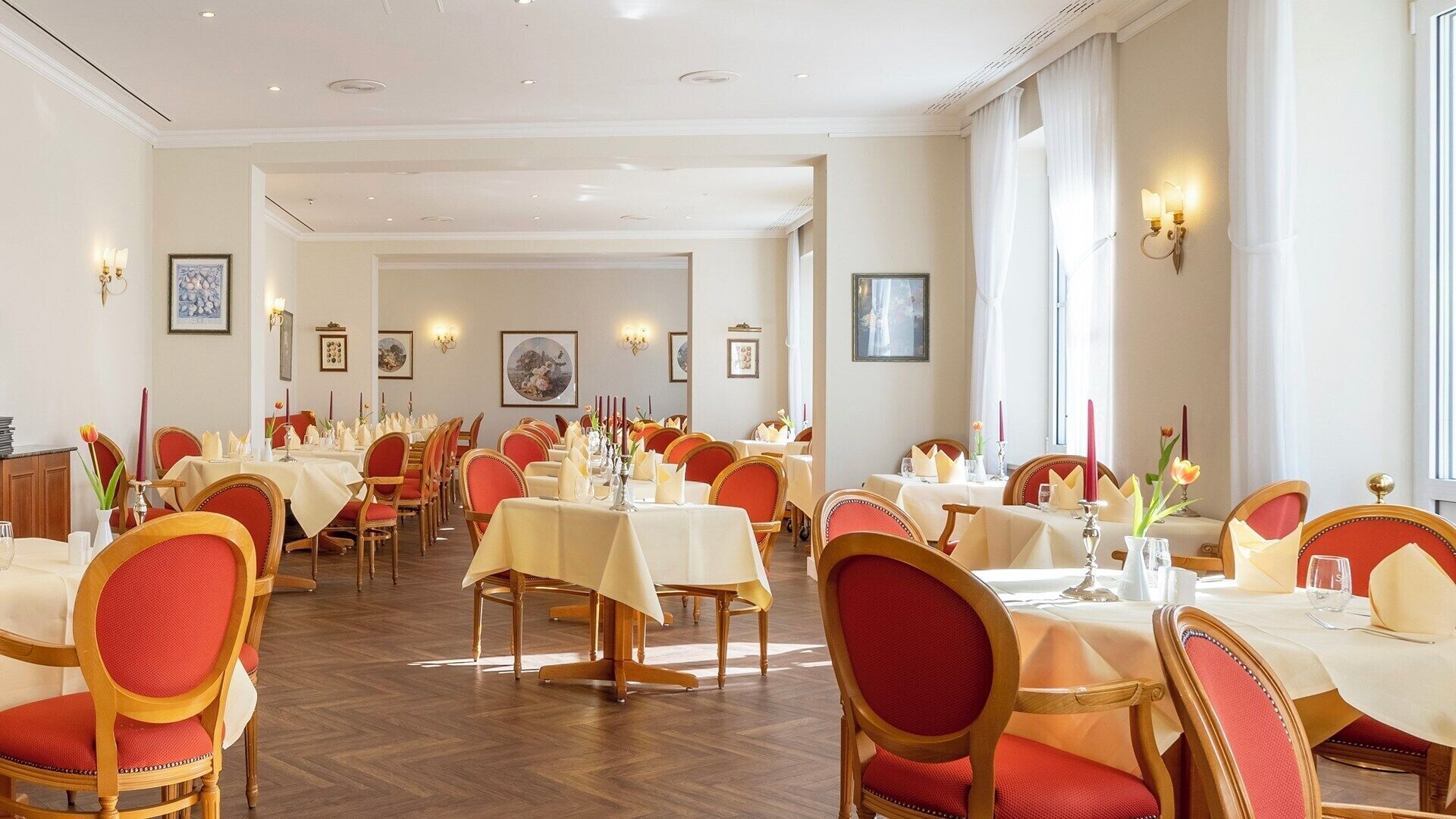 A spacious dining area with multiple tables and chairs, framed artwork on the walls, and a wooden floor.