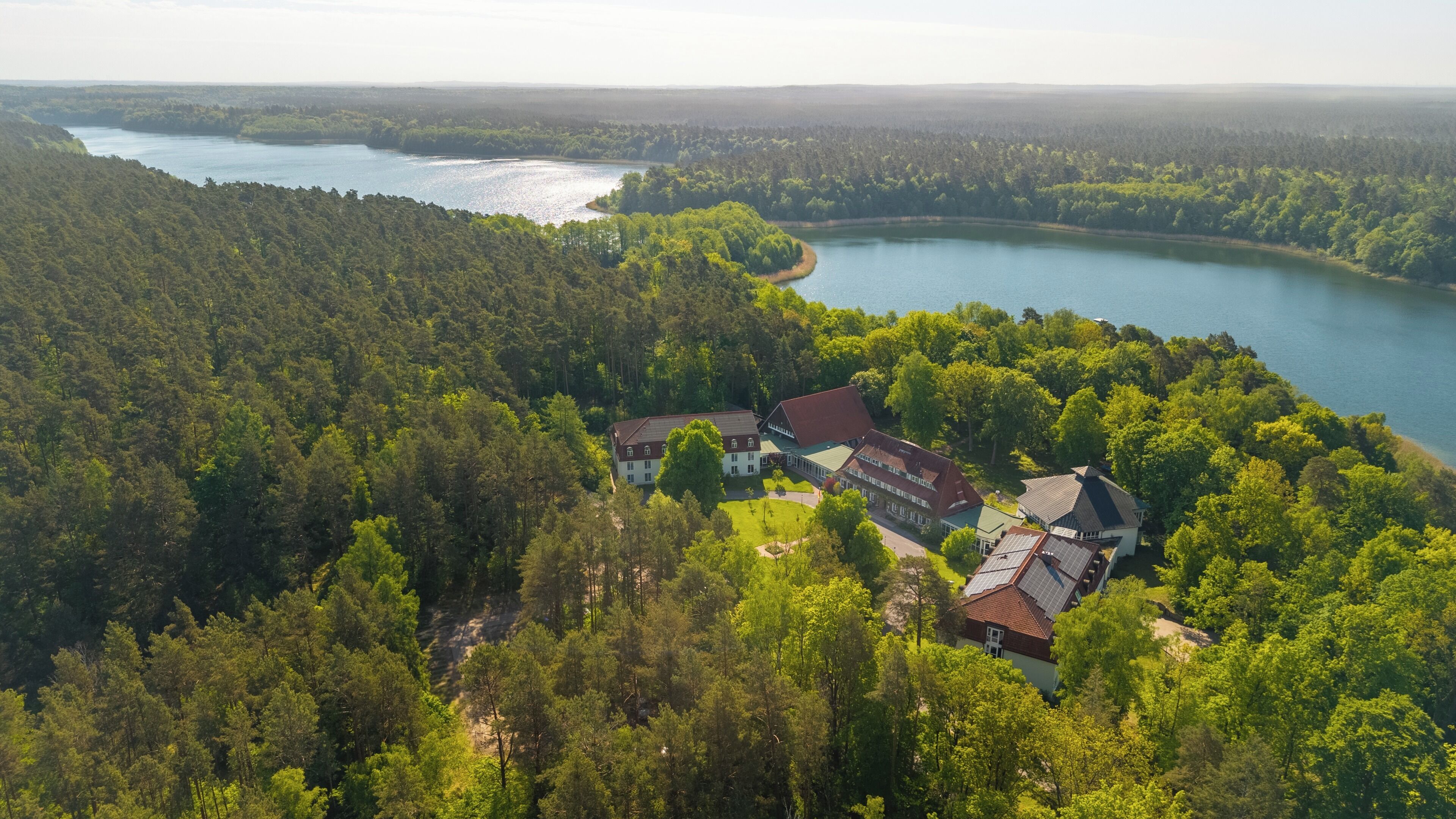 An aerial view of a forested area with a lake and a large building with multiple sections.