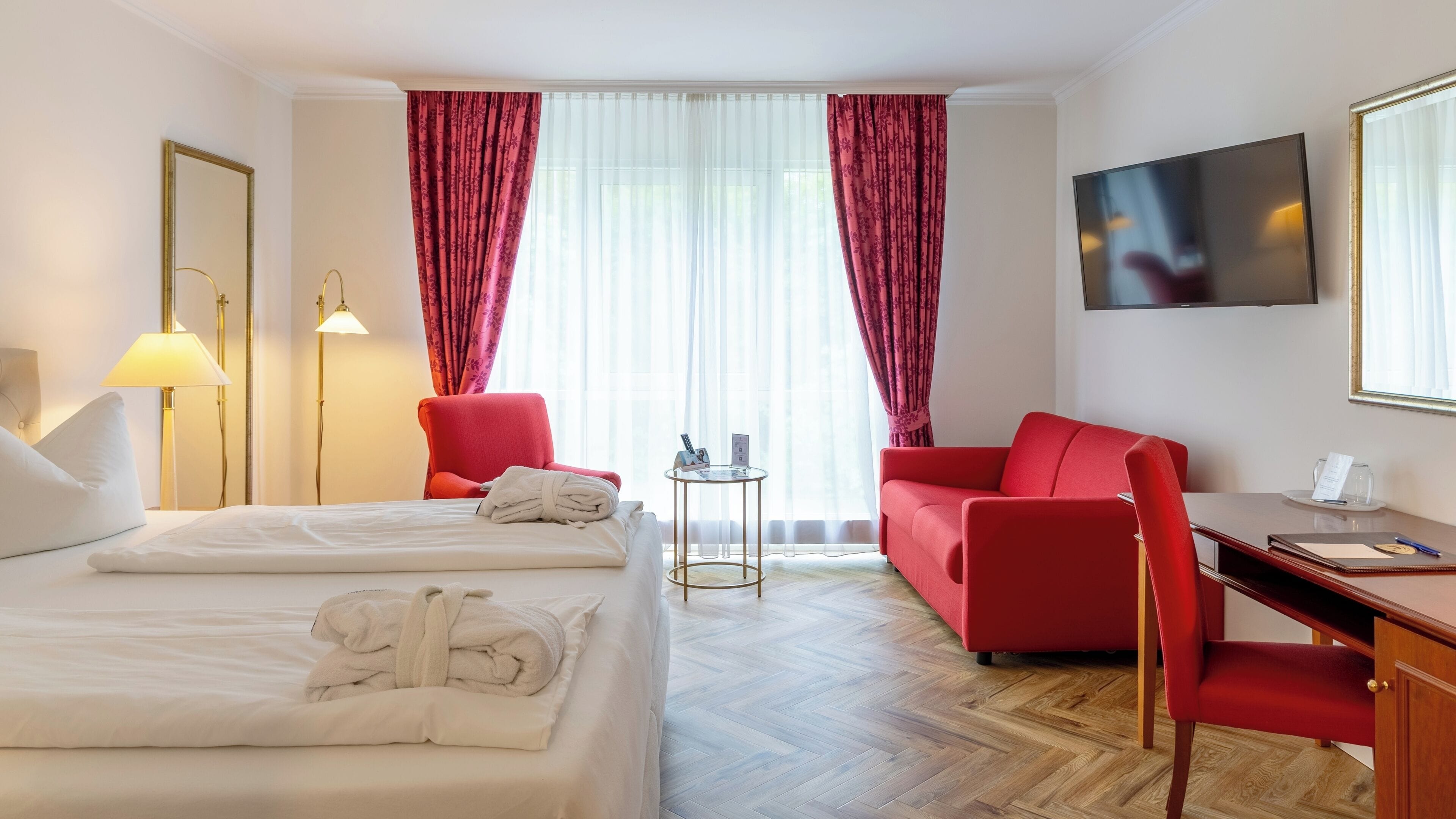 A hotel room with two beds, a desk, a chair, a TV, and a small table with a lamp.