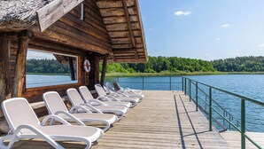 Una terraza de madera con sillas de descanso frente a un lago, un flotador salvavidas y una estructura con techo de paja.