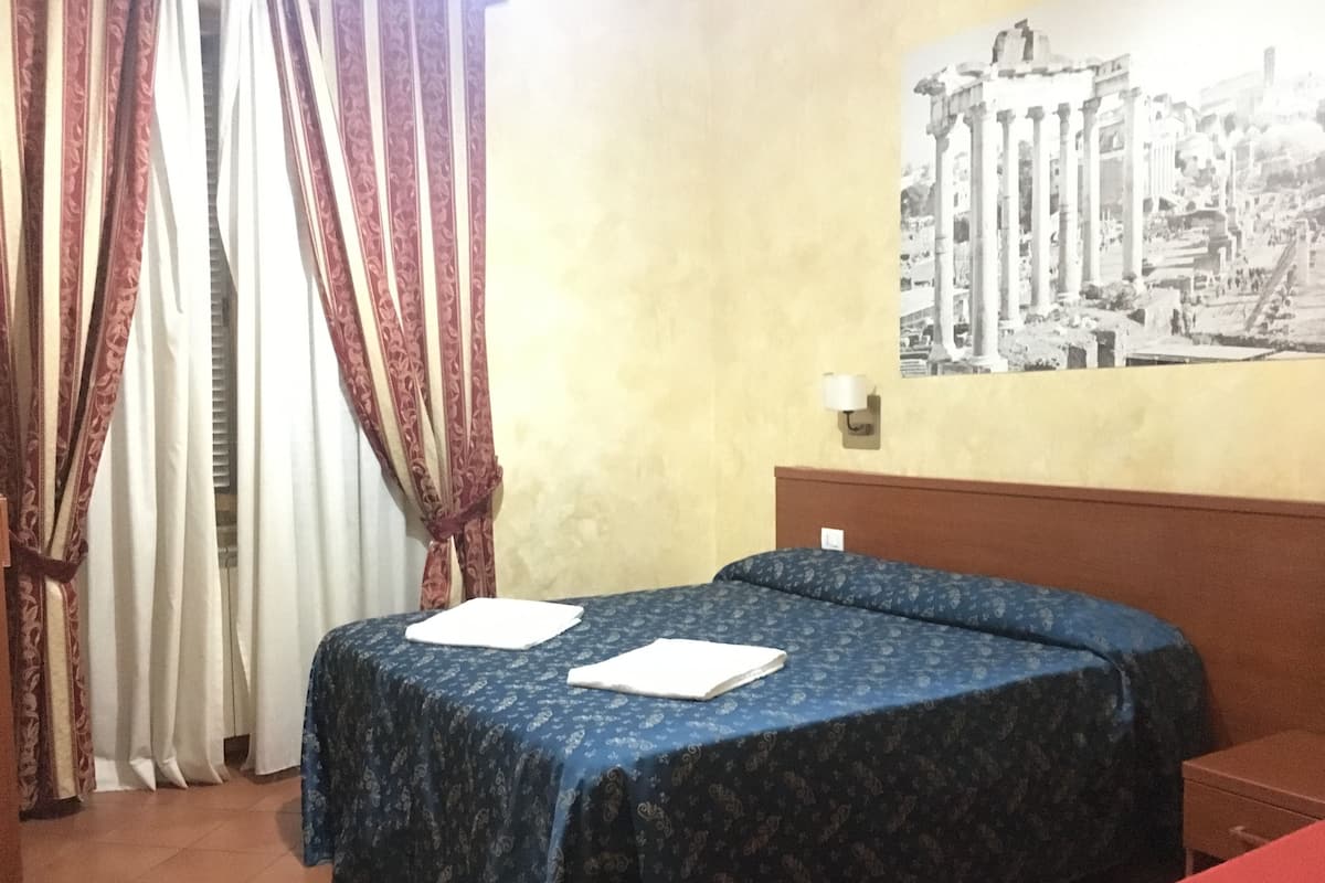 A hotel room with two beds, a red bedspread, a patterned blue bedspread, a wall-mounted lamp, and a black and white photo of ancient ruins.