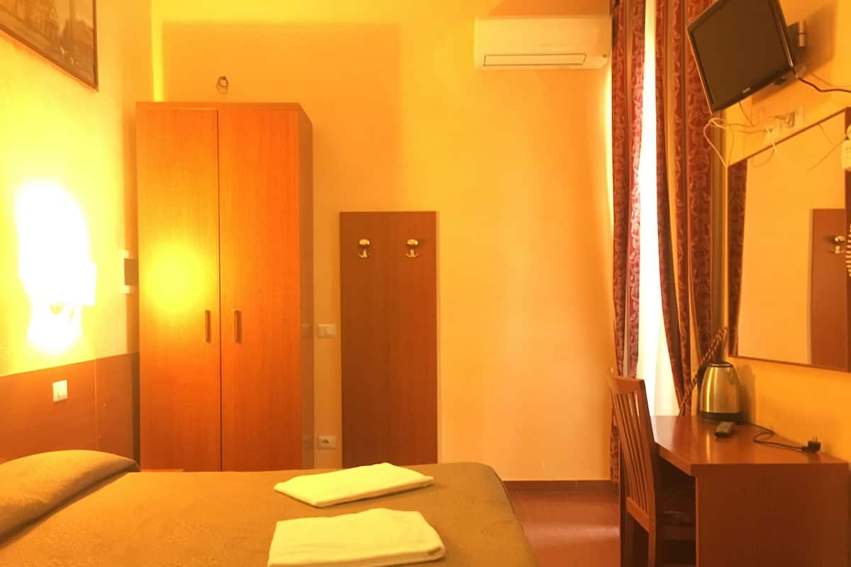 A hotel room with a bed, a wardrobe, a desk, and an air conditioning unit.