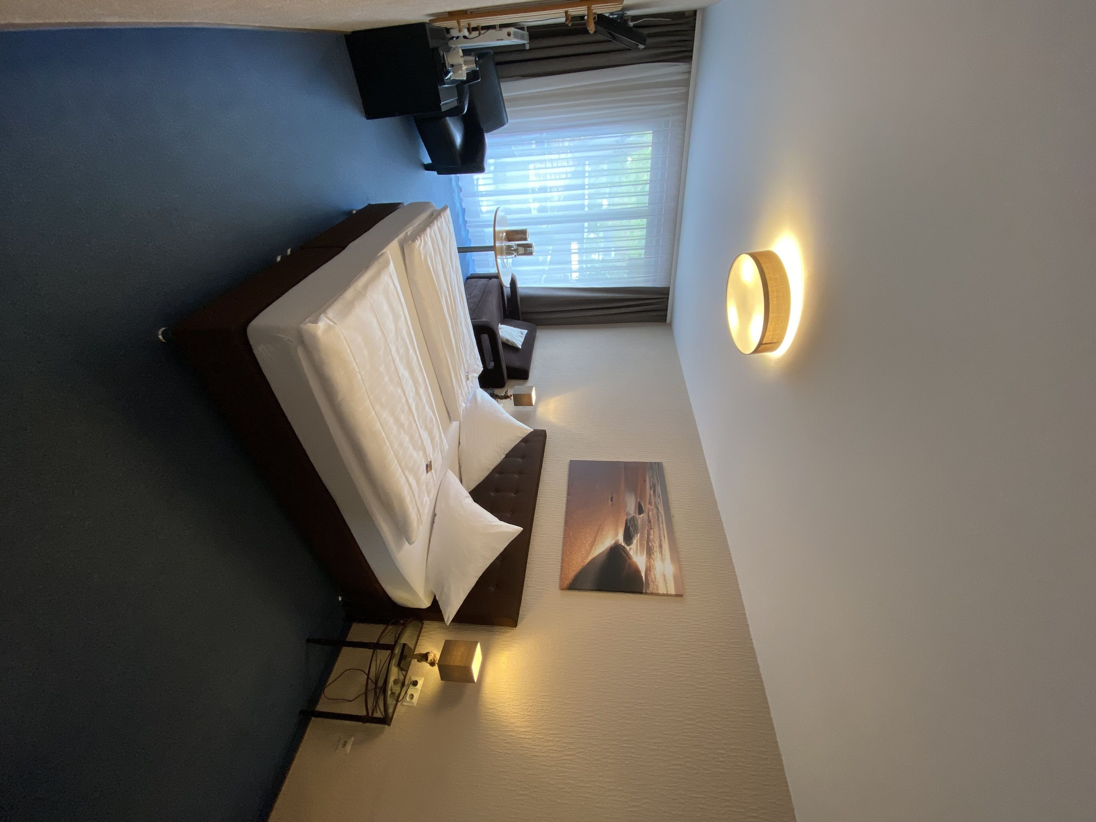 A hotel room with a bed, a small table, a chair, a TV, and a window with blinds.