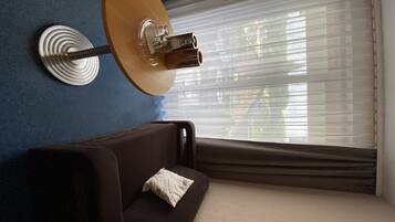 Ein Zimmer mit einer braunen Couch, einem weißen Kissen, einer Wandlampe und einem Fenster mit Jalousien.
