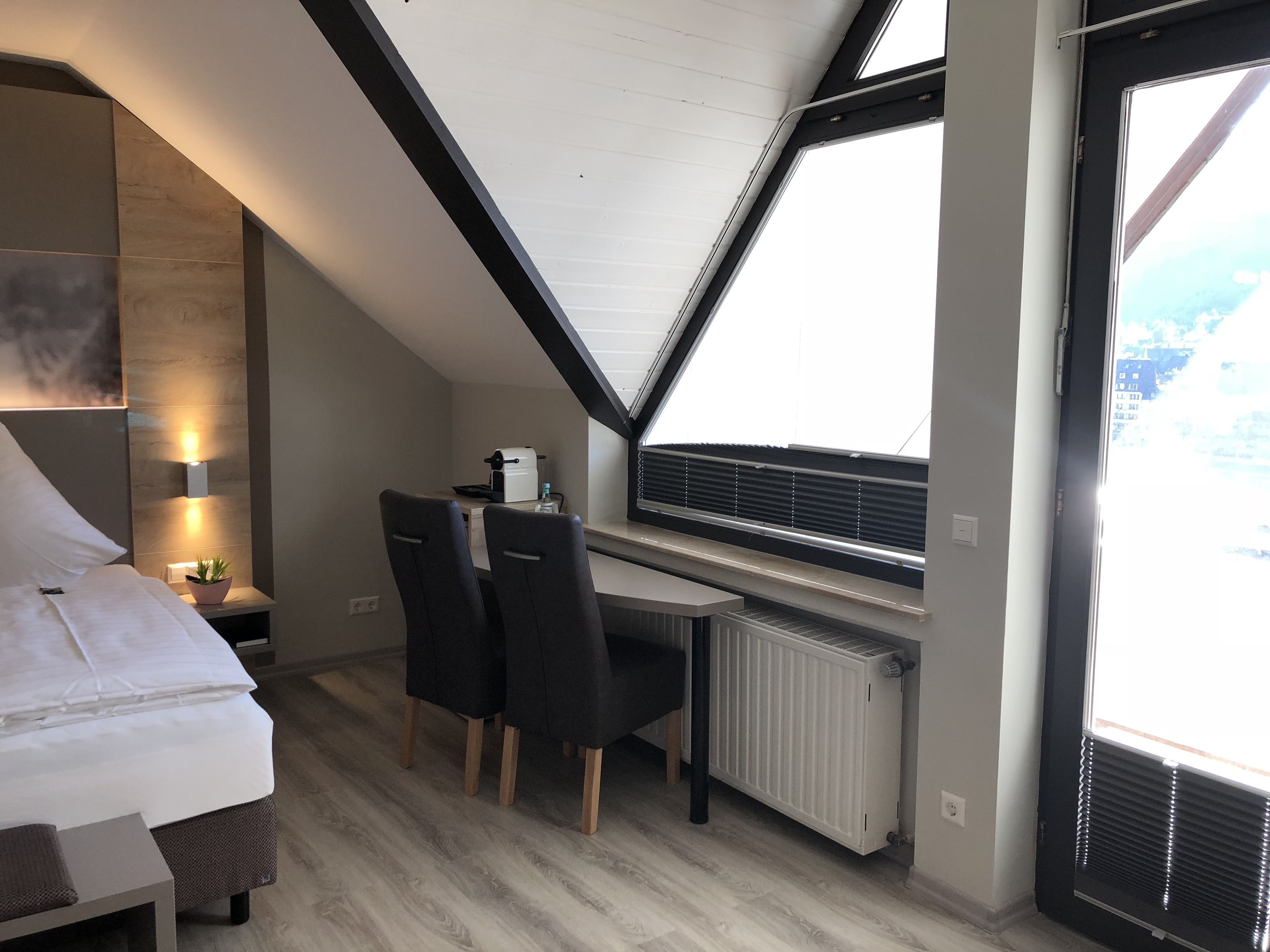 Een moderne slaapkamer met een schuine plafond, een bed, een bureau met twee stoelen, een radiator en een raam met jaloezieën.