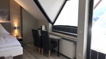 Een moderne slaapkamer met een schuine plafond, een bed, een bureau met twee stoelen, een radiator en een raam met jaloezieën.