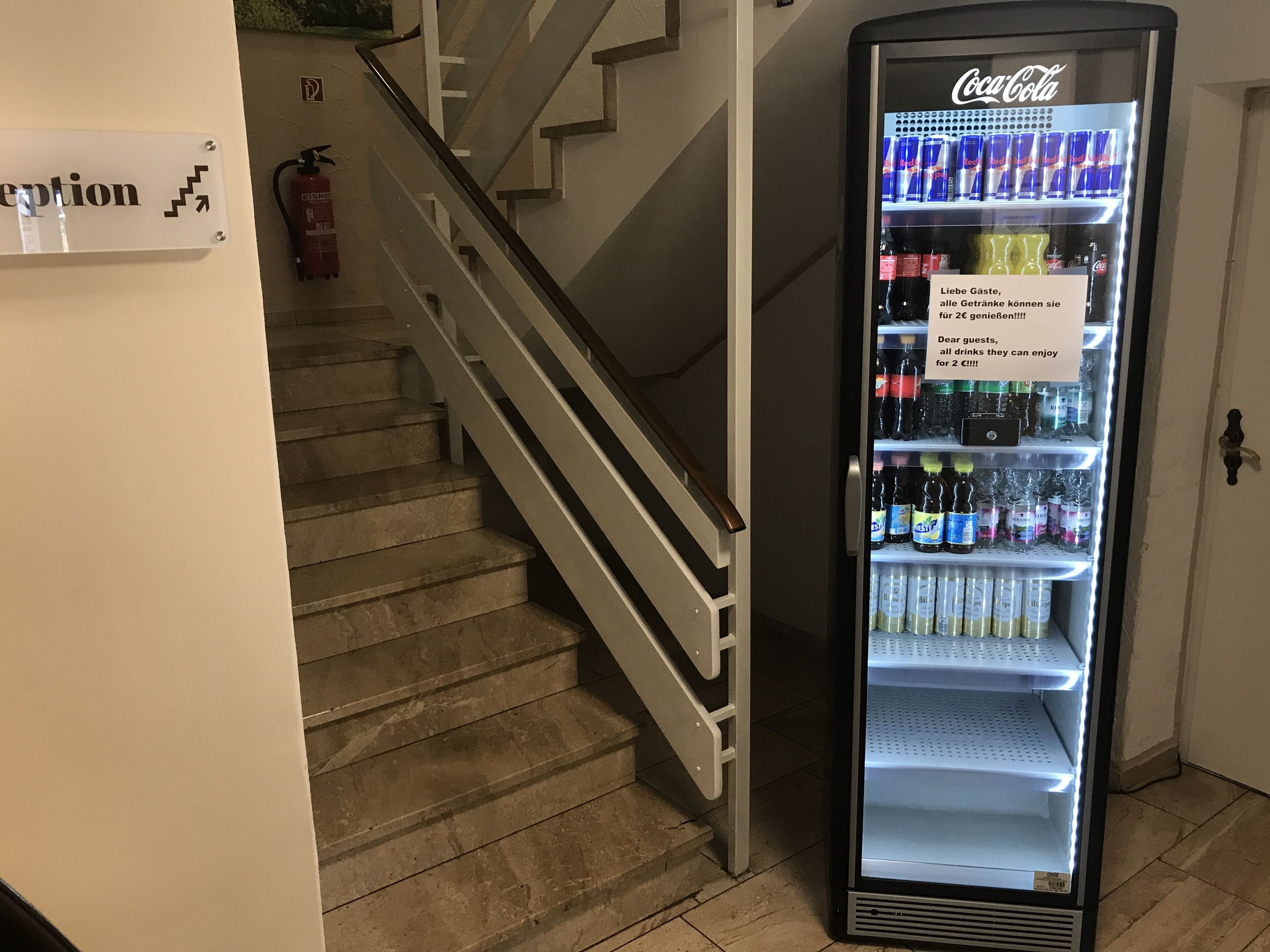 A Coca-Cola refrigerator stocked with beverages, a staircase, and a fire extinguisher.