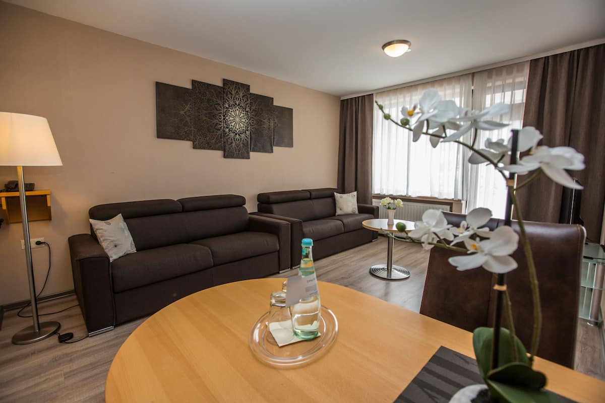 Una sala moderna con un sofá, una mesa de centro y una mesa de comedor. Hay una pieza decorativa en la pared sobre el sofá y un jarrón con flores blancas sobre la mesa de comedor.
