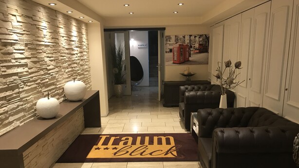 A reception area with a stone wall, a reception desk, a 'Traumblick' mat, leather sofas, and a door leading to a hallway. - Hotel Traumblick (Cochem)
