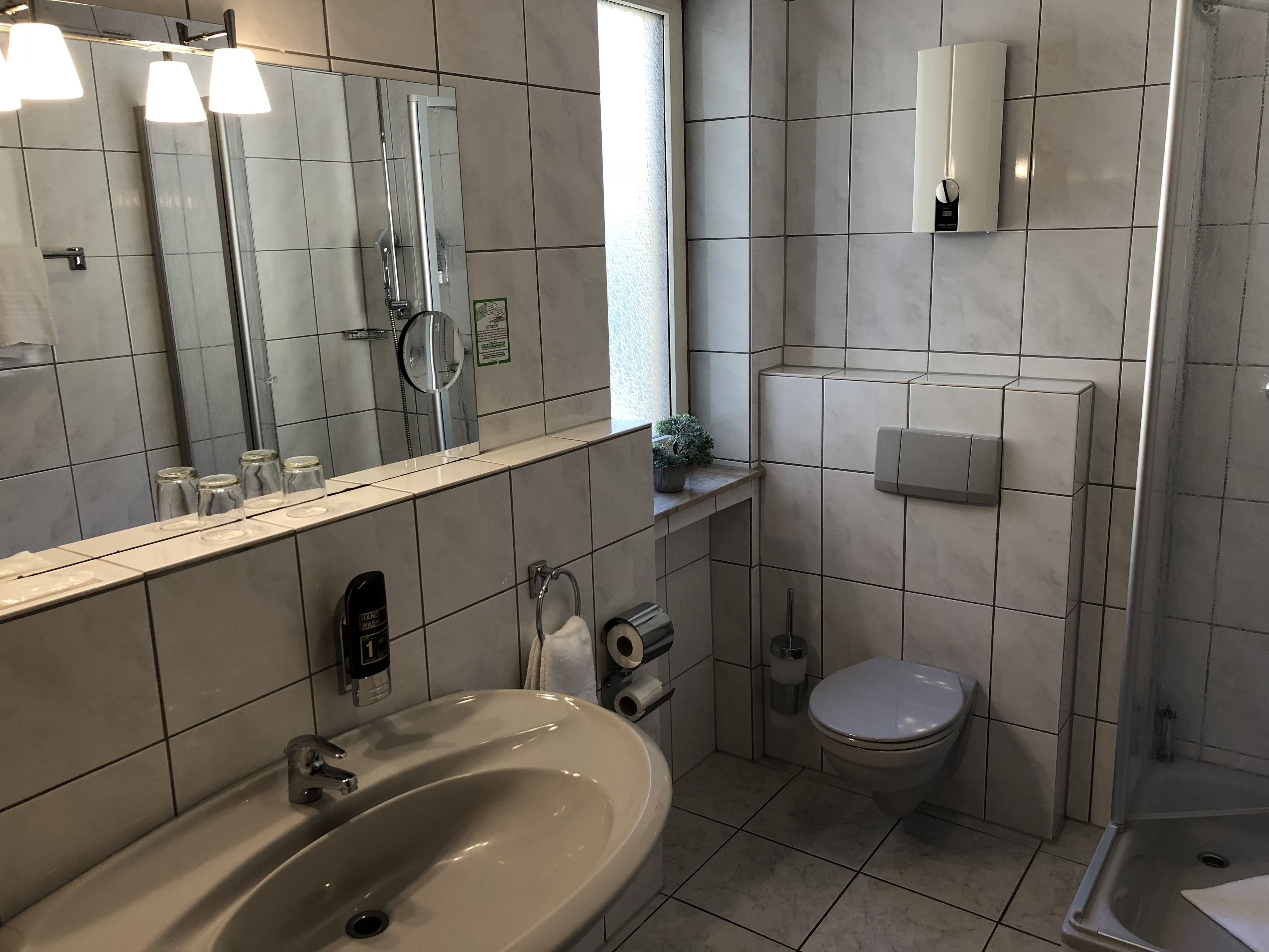 A modern bathroom with a sink, toilet, and shower. It features a large mirror, a towel dispenser, and a small potted plant.