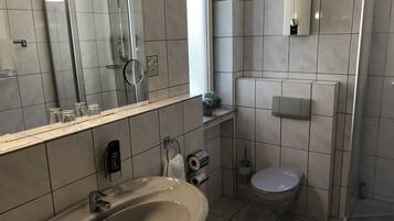 A modern bathroom with a sink, toilet, and shower. It features a large mirror, a towel dispenser, and a small potted plant.