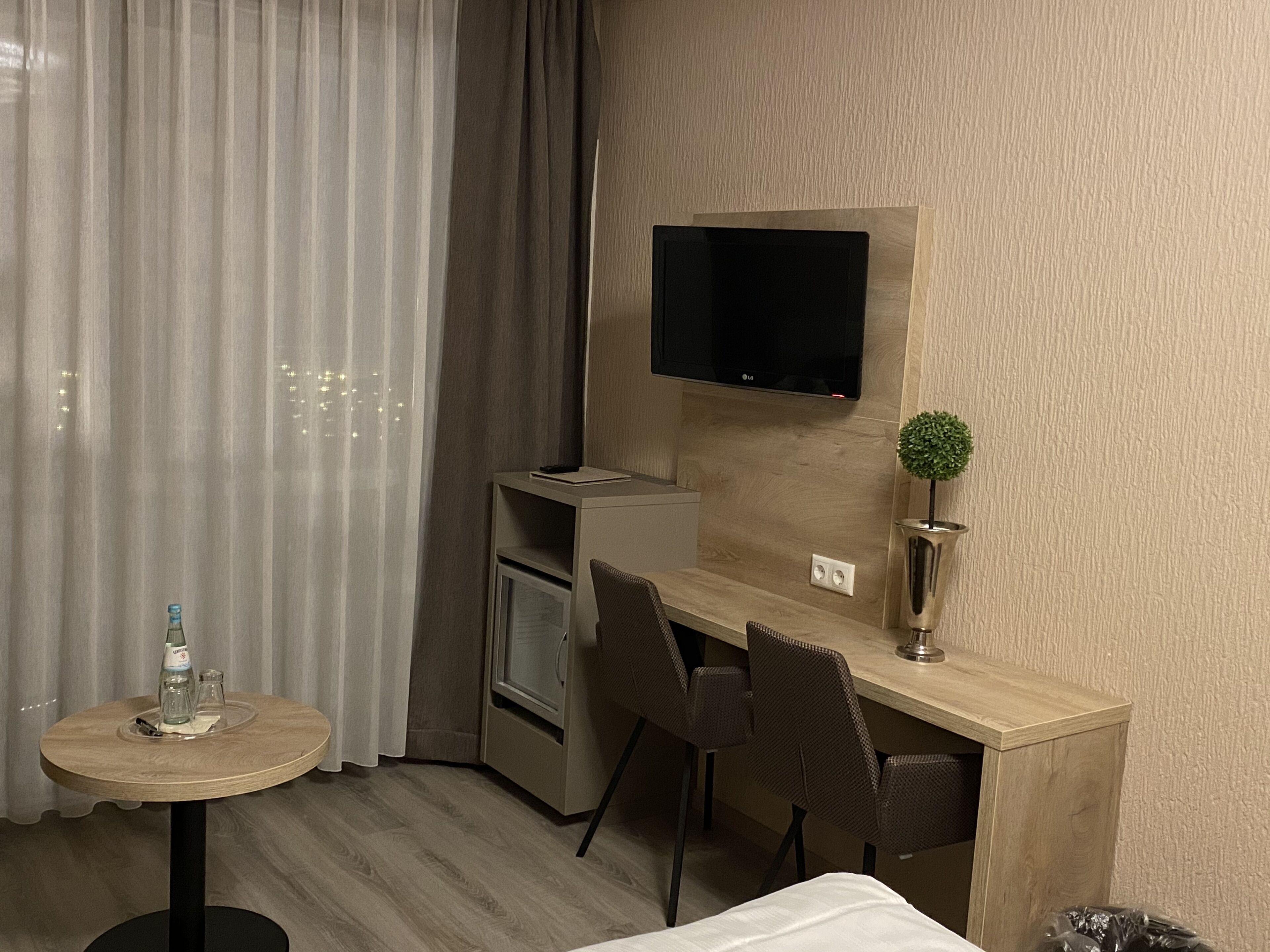 Habitación de hotel con un escritorio de madera, dos sillas, un televisor montado en la pared, una pequeña planta en maceta, una botella de agua y un vaso sobre la mesa.