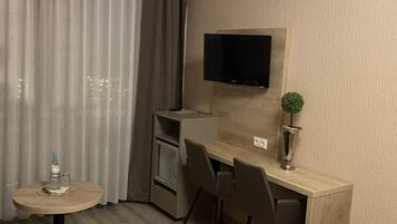Habitación de hotel con un escritorio de madera, dos sillas, un televisor montado en la pared, una pequeña planta en maceta, una botella de agua y un vaso sobre la mesa.
