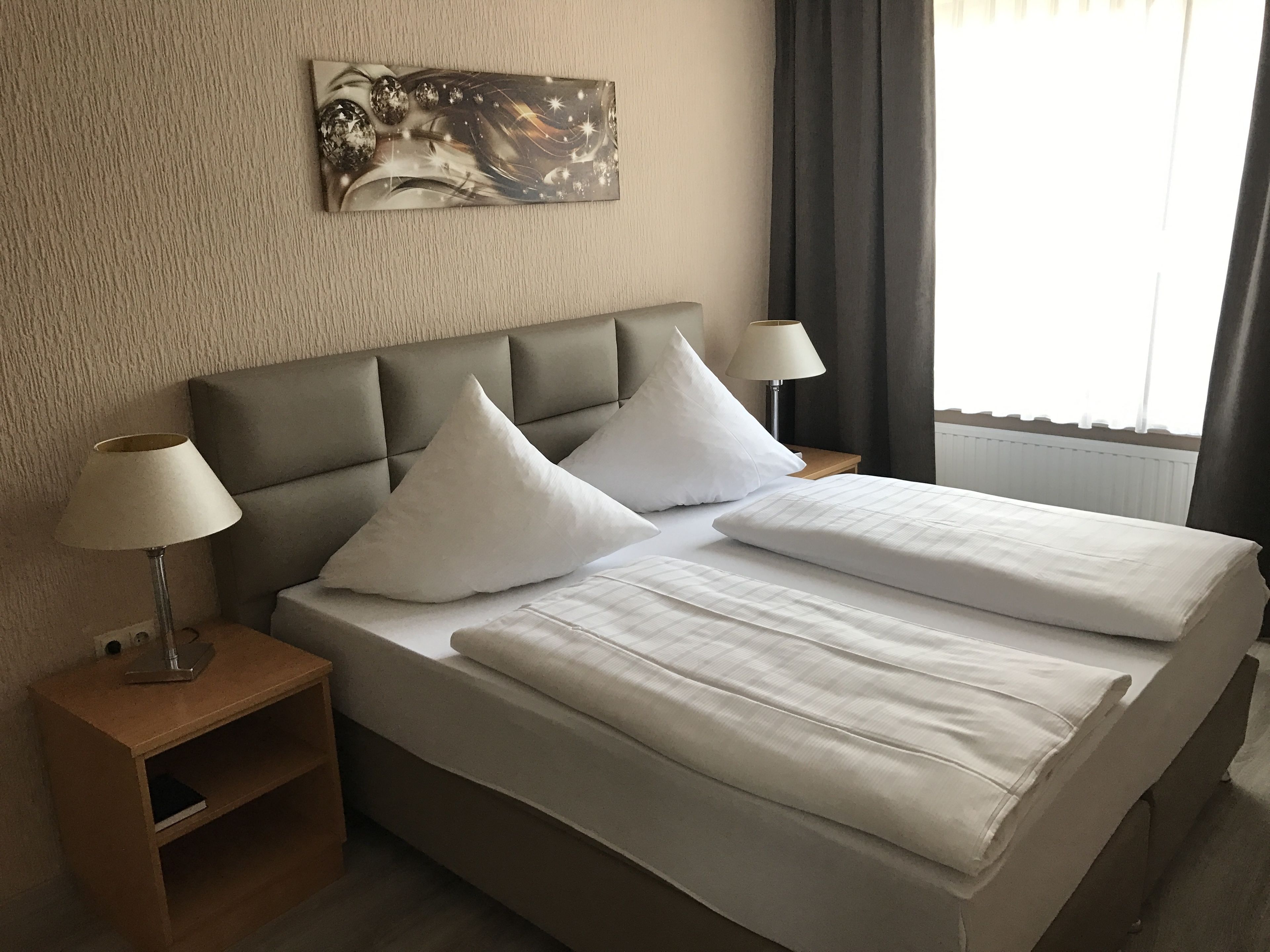 Una cama bien hecha con sábanas blancas, un cabecero, dos lámparas de noche y una mesita de madera con un libro.