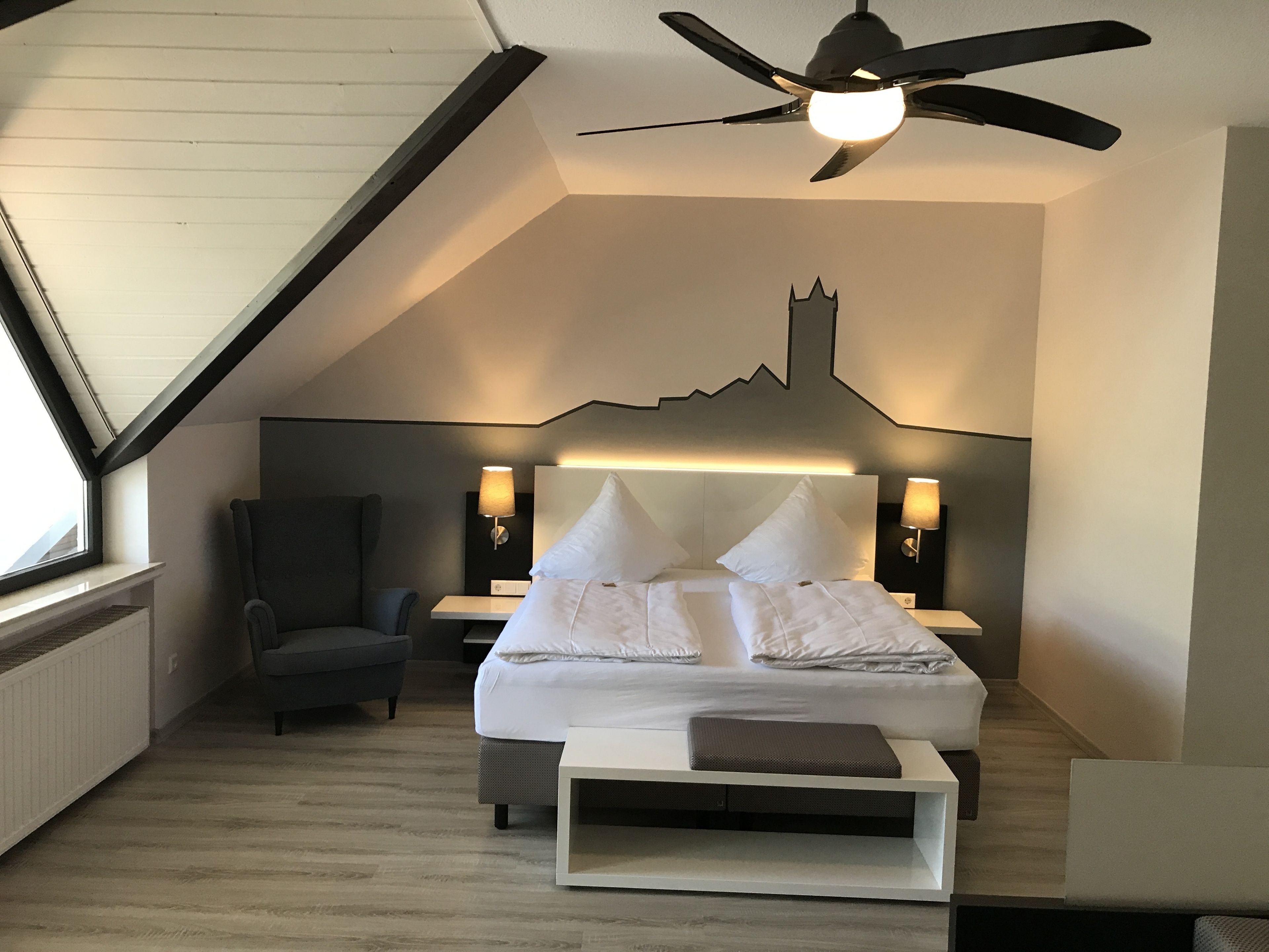 A modern hotel room with a sloped ceiling, a bed with white linens, two bedside lamps, a chair, and a small table.