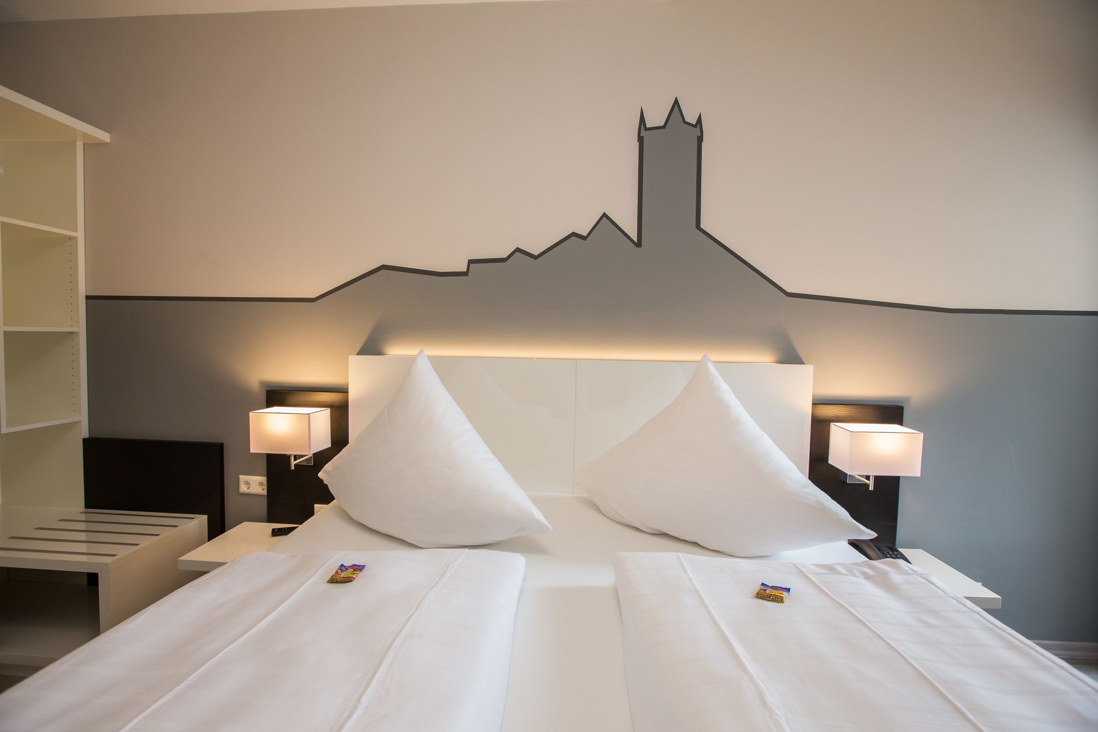 Ein Hotelzimmer mit einem Bett, zwei Nachttischen, zwei Lampen und einem an der Wand hängenden Stadtbild.