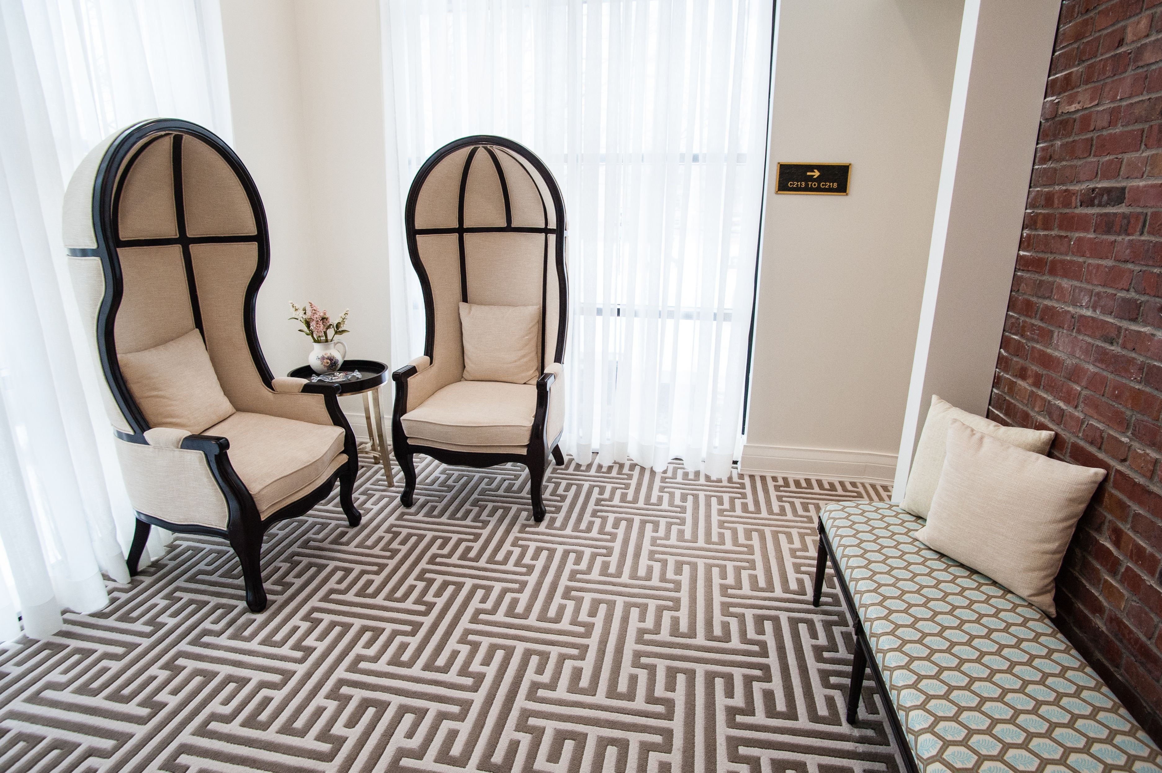 A room with two armchairs, a small table with a vase, a patterned floor, and a bed with pillows.