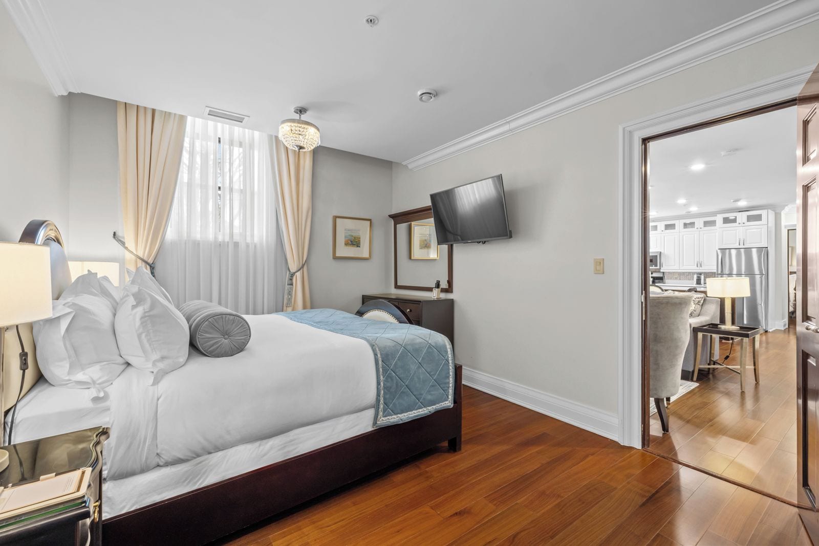 A hotel room with a bed, bedside table, lamp, curtains, a television, and a view into the kitchen area.