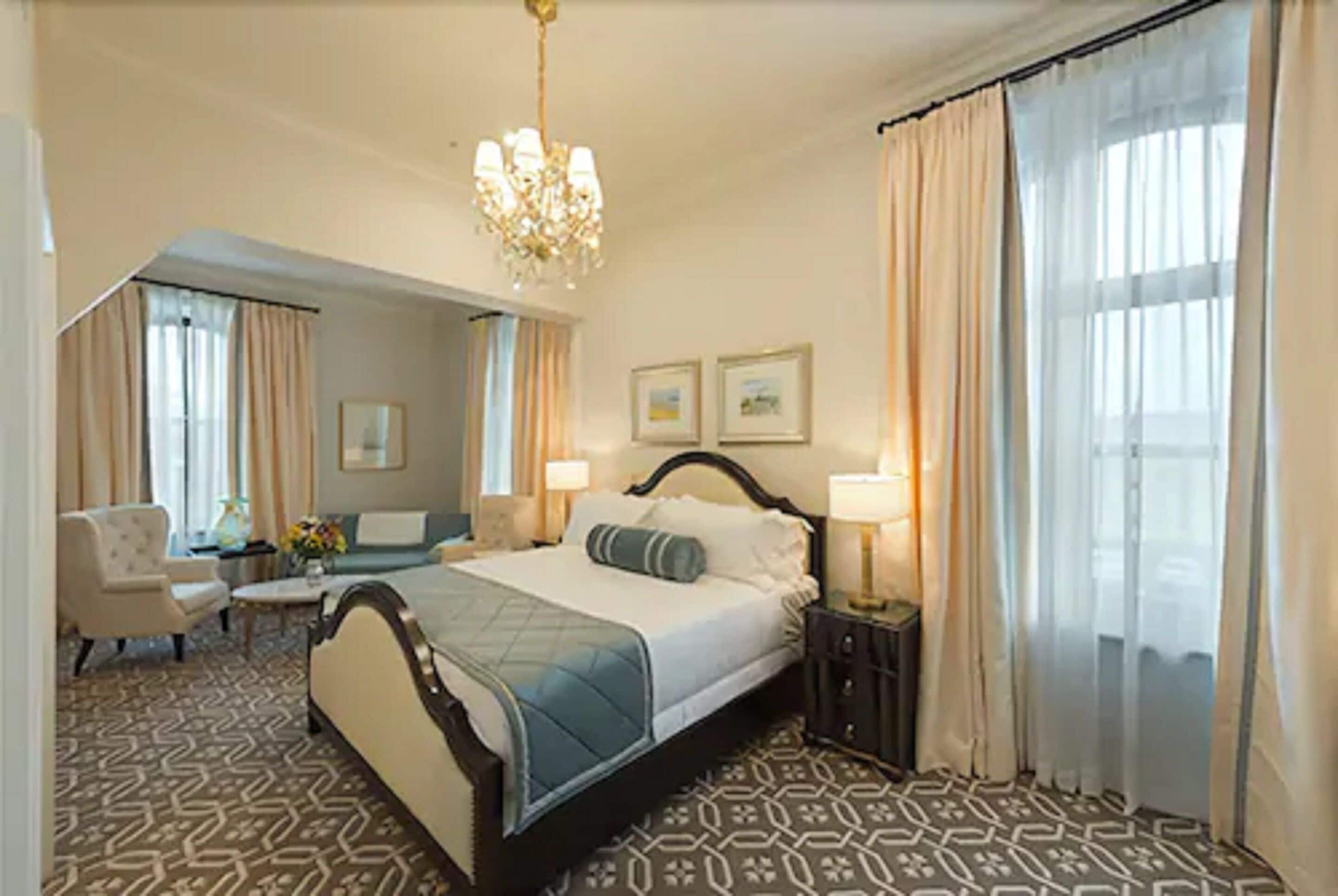 Habitación de hotel con una cama grande, un área de descanso con una silla y una mesa, un candelabro y un ventanal con cortinas.