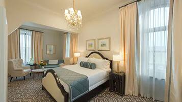 Habitación de hotel con una cama grande, un área de descanso con una silla y una mesa, un candelabro y un ventanal con cortinas.