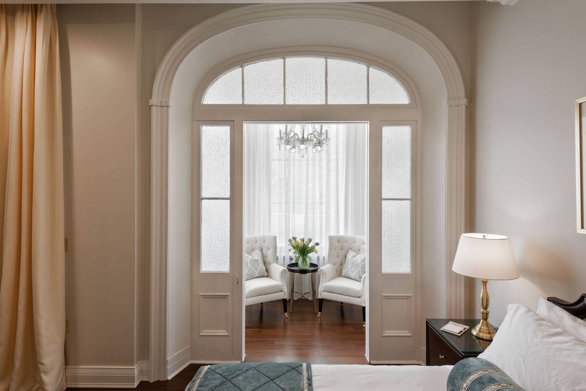 A hotel room with a large arched doorway leading to a sitting area with two chairs and a table, a chandelier, and a bedside lamp.