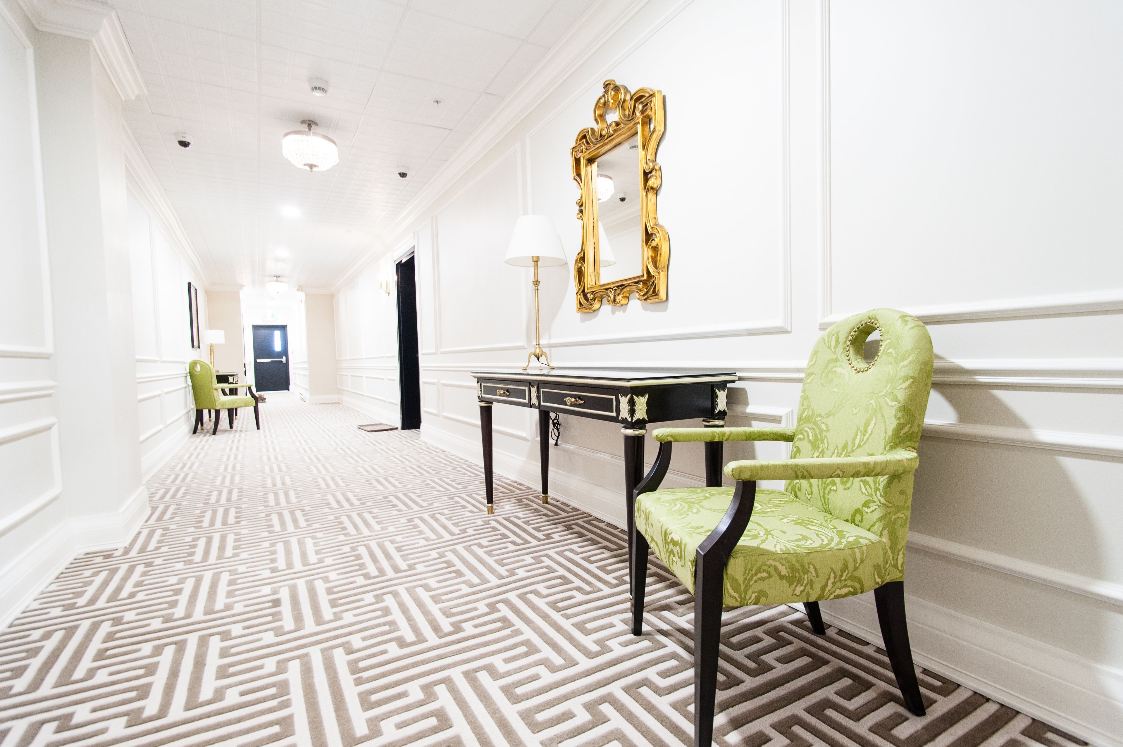 Un couloir avec un tapis à motifs, une chaise verte, une table avec une lampe et un miroir encadré d’or accroché au mur.