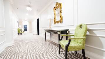 Un pasillo con alfombra estampada, una silla verde, una mesa con lámpara y un espejo con marco dorado en la pared.
